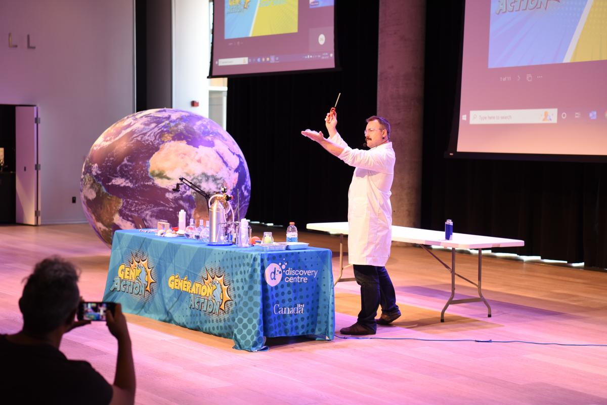 A person in a white coat standing in front of a table with a globe and a projection screen